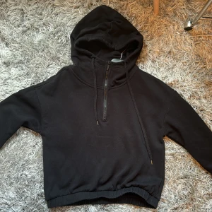 Svart hoodie med dragkedja - Säljer en svart hoodie i bra skick. Den har en halv dragkedja framtill och en justerbar huva med snören. Perfekt för en avslappnad stil och passar till alla tillfällen. Supermjuk och bekväm!