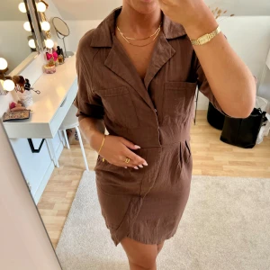 Brun miniklänning - Superfin brun miniklänning i linneliknande material. Storlek 36 men passar även 34. Knäppning i sidan med dragkedja och framtill med knappar. Är 167 cm lång.