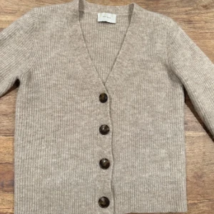 Beige ribbad kofta från Neo Noir - Säljer en stilren beige kofta från Neo Noir. Den är ribbad med en v-ringning och har stora knappar framtill. Perfekt för att hålla sig varm och snygg under kyligare dagar.