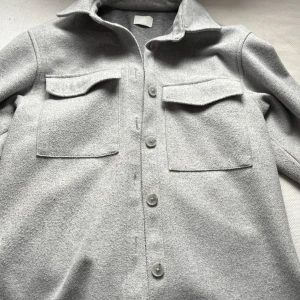 Grå Jacka - Snygg grå jacka framtill och två stora bröstfickor med lock. Perfekt för lager-på-lager-stil. Materialet ser mjukt och bekvämt ut, vilket gör den idealisk för kyligare dagar.