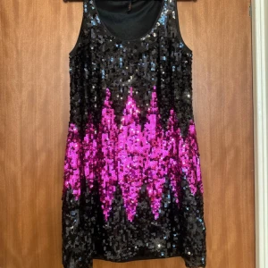 Svart och rosa paljettklänning - Helt ny Snygg svart klänning täckt med glittriga paljetter i ett coolt mönster med rosa detaljer. Perfekt för att sticka ut på festen! Klänningen är ärmlös och har en kort längd.
