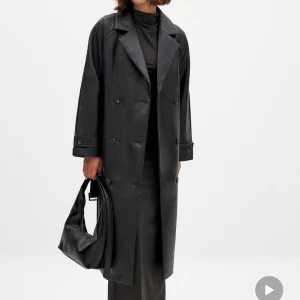 Svart trenchcoat i PU - Snygg svart trenchcoat i PU-material med knappar och bälte i midjan. Den har en klassisk design med långa ärmar och en stilren look. Perfekt för att ge en edgy touch till din outfit. Använd EN gång, känner att den är lite lång på mig, men super snygg oversized look om man har en mindre storlek och är ute efter det