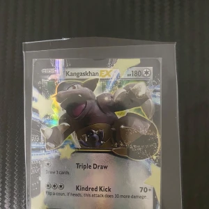 Kangaskhan EX Pokémonkort - Säljer ett Kangaskhan EX Pokémonkort med HP 180. Kortet har en holografisk yta och visar Kangaskhan i en kraftfull pose. Det har två attacker: Triple Draw och Kindred Kick. Perfekt för samlare eller spelare som vill förstärka sin kortlek.