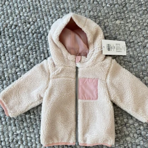 Beige teddyjacka från Name It - Säljer en supermjuk och gosig beige teddyjacka från Name It. Jackan har en söt rosa ficka på bröstet och dragkedja framtill. Perfekt för att hålla de små varma och stiliga. 🧸Storlek 62.