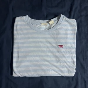 Randig t-shirt från Levi's - Säljer en stilren ljusblå och vit randig t-shirt från Levi's. Den har en klassisk rund halsringning och korta ärmar. Perfekt för en avslappnad look.