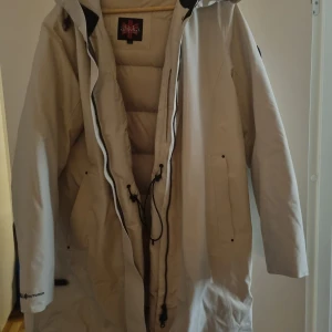 Beige dunjacka från áhkká - Beige dunjacka från áhkká med huva och dragkedja. Jackan har långa ärmar och är perfekt för kyligare väder. Den har praktiska fickor och justerbar dragsko i midjan.
