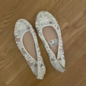 Vita blommiga ballerinaskor - Säljer ett par vita ballerinaskor med blommigt mönster och spetsdetaljer. Skorna har en rund tå och är i storlek dam 35. Perfekta för en somrig look! Inga defekter