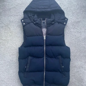 Mörkblå dunväst med huva - Säljer en snygg mörkblå dunväst med dragkedja och avtagbar huva. Västen har en quiltad designl. Perfekt för kyligare dagar!