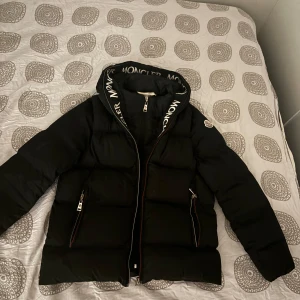 Svart dunjacka från Moncler - Snygg svart dunjacka från Moncler med dragkedja och logotyp på huvan. Jackan har en pufferdesign och långa ärmar, perfekt för kyliga dagar. Den har också praktiska fickor med dragkedjor. Dragkedjan på fickorna är trasig därav priset. Strl xs/junior 15-16