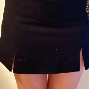 Svart kortkjol med broderad detalj - Svart minikjol med slitsar och guldbroderi framtill, mycket bra skick, jättesnygg men har tyvärr inte kommit till användning😊