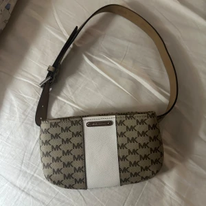 Beige och vit axelväska från Michael Kors - Snygg axelväska från Michael Kors i beige med vitt band och logotypmönster. Väskan har en justerbar axelrem och en dragkedja för säker förvaring. Perfekt för att bära dina nödvändigheter med stil.