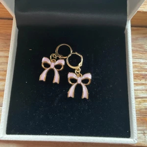 Guldörhängen med rosa rosett - Charmiga örhängen i guld med en söt rosa rosett. Perfekta för att ge en lekfull touch till din outfit. De hänger fint och är lätta att bära hela dagen.