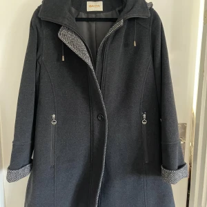 Mörkgrå kappa från Hettemarks - Elegant mörkgrå kappa från Hettemarks i ull och kashmir. Den har en huva och långa ärmar med uppvikta muddar. Kappan stängs med dragkedja och har fickor som stängs med dragkedja framtill. Perfekt för kyliga dagar.