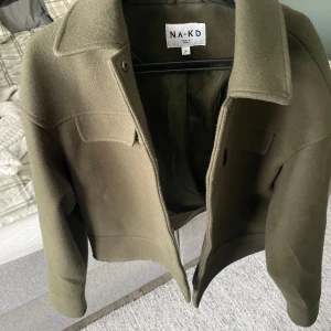 Olivgrön jacka från NA-KD - Säljer en stilren olivgrön jacka från NA-KD. Jackan har en klassisk krage och stängs med knappar framtill. Den har långa ärmar och två bröstfickor som ger en cool look. Perfekt för höst och vår!
