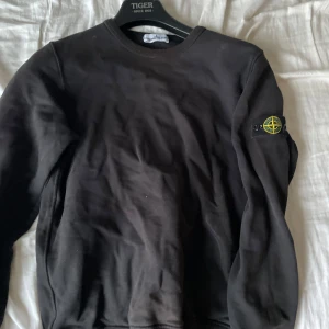 Svart tröja från Stone Island - Säljer en svart tröja från Stone Island med deras ikoniska märke på ärmen. Tröjan har en klassisk rund hals och långa ärmar. Perfekt för en stilren och avslappnad look.  Tröjan är största barn storlek och sitter som XS/S