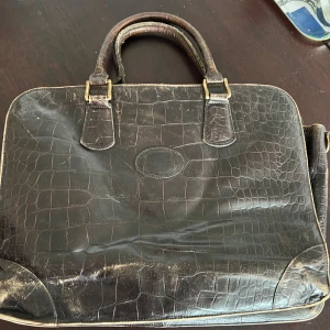 Svart skinnportfölj från mulberry med krokodilmönster - Elegant svart portfölj i skinn med krokodilmönster. Väskan har två handtag och en dragkedja upptill. Insidan är fodrad med ett rutigt tyg i flera färger. Perfekt för att bära dokument och laptop med stil.