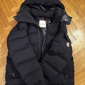 Svart dunjacka från Moncler - Säljer en svart dunjacka från Moncler med huva och dragkedja. Jackan har långa ärmar och två fickor med dragkedja. Perfekt för kalla dagar med sin pufferdesign. Tar emot byte