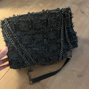 Svart axelväska  - Snygg svart axelväska med fransiga detaljer och kedjeaxelrem. Väskan har en praktisk innerficka med dragkedja och stängs med en tryckknapp. Den är rymlig och stor🤩använd en gång