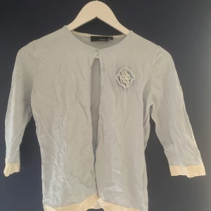 Ljusblå kofta från Hennes - Säljer en ljusblå kofta från Hennes med trekvartsärmar och en dekorativ blomdetalj på bröstet. Koftan har en rund halsringning och är tillverkad i en mjuk blandning av viskos och polyamid. Perfekt för en stilren look.