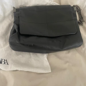 Svart axelväska med kedja - Snygg svart axelväska från Zara i skinn med en stilren design. Väskan har en kedja som axelrem och flera fack med dragkedja för praktisk förvaring. Säljer då den inte används längre