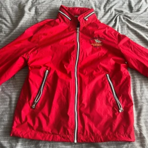 Röd vindjacka från East West - Säljer en snygg röd vindjacka från East West med broderad text på bröstet. Jackan har dragkedja framtill och två fickor med dragkedjor. Perfekt för blåsiga dagar och har en sportig look.
