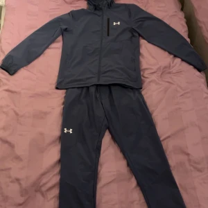 Mörkblå träningsset från Under Armour - Säljer ett mörkblått träningsset från Under Armour. Setet består av en jacka med dragkedja och matchande byxor. Perfekt för träning eller avslappnade dagar. Jackan har en liten logotyp på bröstet och byxorna har en logotyp på benet. Tracksuiten säljs inte längre den är köpt på jd sports för 2450kr jackan är storlek M, byxan S men funkar som M också