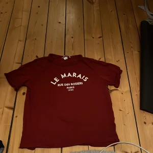 Röd t-shirt med tryck - Säljer en röd t-shirt från H&M med texten 'LE MARAIS RUE DES ROSIERS PARIS 1985' tryckt på framsidan. T-shirten har en avslappnad passform och korta ärmar. Perfekt för en casual look.