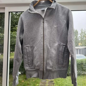 Grå fleecejacka från Nike - Säljer en grå fleecejacka från Nike med dragkedja och ribbade muddar. Jackan har långa ärmar och två fickor framtill. Perfekt för kyliga dagar.