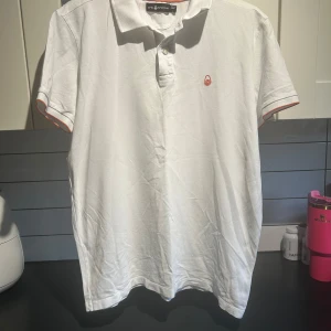 Vit pikétröja från Sail Racing - Säljer en stilren vit pikétröja från Sail Racing. Tröjan har korta ärmar och en klassisk krage med knappar. Den har en liten logotyp broderad på bröstet och orange detaljer vid ärmslut och krage. Perfekt för en avslappnad och sportig look.