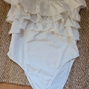 Vit volangbody - Säljer en stilren vit body med flera lager volanger upptill. Perfekt för en elegant look. Den är axelbandslös och har en tight passform som framhäver figuren.