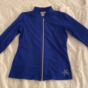 Blå jacka från K-Skate - Snygg blå jacka från K-Skate med dragkedja och glittrande detaljer. Perfekt för konståkning eller andra aktiviteter. Jackan har långa ärmar och en bekväm passform.