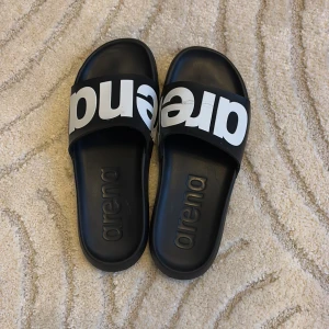 Svarta badsandaler från Arena - Snygga svarta badsandaler från Arena med stor vit logotyp över remmen. Perfekta för stranden eller poolen.