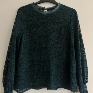 Mörkgrön spetsblus från Happy Holly - Elegant mörkgrön spetsblus från Happy Holly med långa ärmar och ett vackert blommönster. Perfekt för att ge en sofistikerad touch till din outfit. Blusen har en rund halsringning och en liten knappdetalj i nacken.