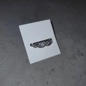 Silverfärgad ring med flätad design - Elegant silverfärgad ring med en unik flätad design. Perfekt för att ge en stilren touch till din look. Ringen har en justerbar öppning för att passa olika storlekar.