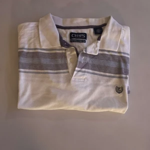 Vit och grå randig pikétröja från Chaps - Säljer en stilren vit och grå randig pikétröja från Chaps. Tröjan har en klassisk krage och knappar framtill. Den är dekorerad med ett broderat emblem på bröstet. Perfekt för en avslappnad och snygg look. 