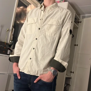 C.P. Company overshirt  - Säljer en stilren beige overshirt från C.P. Company. Overshirten har långa ärmar och knappar framtill samt på ärmsluten. Den är tillverkad i ett lätt och bekvämt material, perfekt för en avslappnad look. Märkets ikoniska linsdetalj finns på ärmen. overshirten är i storlek L men passar så här på mig som är 181 cm lång och väger 67 kg hetsbevis finns genom cirtilogo och nypris ligger runt 3000 Overshirten är i toppskick utan några defekter.  Byten är intressant 