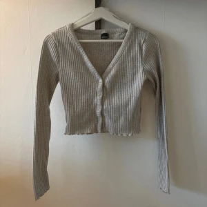 Grå ribbad kofta från Gina Tricot - Säljer en stilren grå ribbad kofta från Gina Tricot. Den har en v-ringad design med knappar framtill och är croppad för en modern look. Perfekt för lager-på-lager under kyligare dagar.