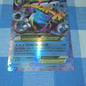 Mega Blastoise-EX Pokémonkort - Säljer ett Mega Blastoise-EX Pokémonkort med holografisk design. Kortet har en kraftfull attack som heter Hydro Bombard och är en del av en samling från 2016. Perfekt för samlare eller spelare som vill stärka sin kortlek. Den har repor på baksidan och så är det lite vit. Såklart är den riktig