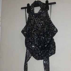 Svart glittrig halterneck topp - Snygg svart halterneck topp med glittriga paljetter över hela ytan. Toppen har en knytning i ryggen och är perfekt för en festlig kväll. Den är ärmlös och har en elegant design som verkligen sticker ut.