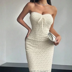 Champagnefärgad spetsklänning - Elegant axelbandslös klänning i champagnefärgad spets med knytning framtill. Perfekt för en stilfull kväll med sin figurnära passform och långa längd. Klänningen har en subtil slits som ger extra rörelsefrihet.