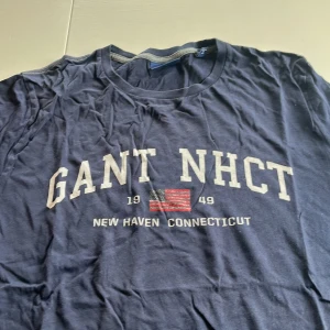 Blå t-shirt från GANT - Säljer en blå t-shirt från GANT med tryck 'GANT NHCT' och en amerikansk flagga. Texten 'New Haven Connecticut' finns också på framsidan. Perfekt för en avslappnad stil.