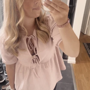 Rosa blus med knytning - Söt superfin rosa blus med knytning framtill och korta ärmar. Perfekt för en avslappnad och stilren look. Blusen har en lätt volangdetalj vid ärmslut och nederkant.