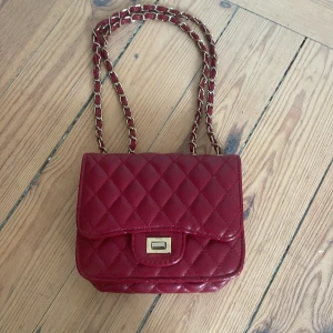 Röd  väska - Snygg röd quiltad axelväska med kedjedetaljer. Väskan har en praktisk dragkedja inuti och en elegant låsdetalj på framsidan. Perfekt för att ge en färgklick till din outfit.