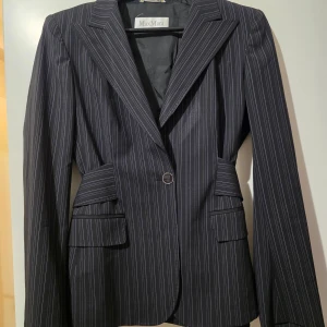 Svart randig kavaj från MaxMara - Elegant svart kavaj från MaxMara med tunna vita ränder. Kavajen har en klassisk design med knappar framtill och fickdetaljer. Tillverkad i Italien, vilket ger en känsla av lyx och kvalitet. Perfekt för en stilren look.