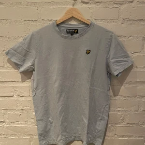 Ljusblå t-shirt från Lyle & Scott - Säljer en stilren ljusblå t-shirt från Lyle & Scott med deras ikoniska logga på bröstet. Perfekt för en avslappnad look. T-shirten har en klassisk passform och korta ärmar.