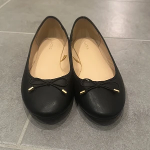 Svarta ballerinaskor med rosett - Eleganta svarta ballerinaskor från Dapors med en söt rosett på ovansidan. Skorna har en klassisk design och är perfekta för en stilren look. De är tillverkade i syntetmaterial och har en bekväm passform. Använt 1 gång.