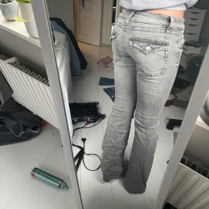 Grå bootcut lågmidjade jeans - Snygga grå bootcut jeans med låg midja och klassisk femficksdesign. Jeansen har en stilren look med dekorativa sömmar och knappar på bakfickorna. Perfekta för en avslappnad stil.