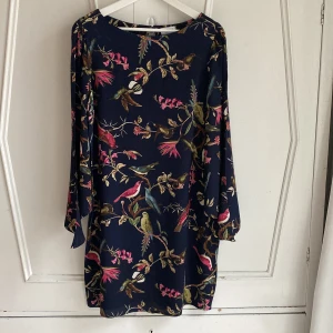 Mönstrad klänning från Vintage Stories - En vacker marinblå klänning från Vintage Stories med ett färgglatt blommönster med fåglar. Klänningen har långa trumpetärmar med slits som kan knytas om man föredrar det. Längd 90. 