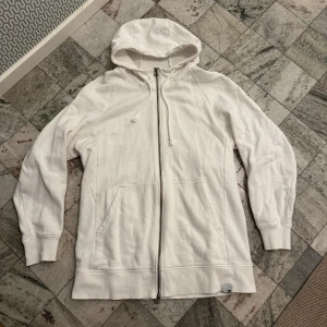Vit hoodie från Adidas - Säljer en stilren vit hoodie från Adidas med dragkedja och huva. Tröjan har långa ärmar och en diskret svart detalj på ryggen. Perfekt för en avslappnad stil.