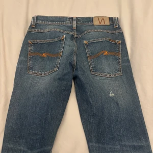 Blå jeans från Nudie Jeans - Snygga blå jeans från Nudie Jeans med slitna detaljer på framsidan och orange sömmar. Klassisk femficksdesign och en bekväm passform. Perfekta för en avslappnad stil.